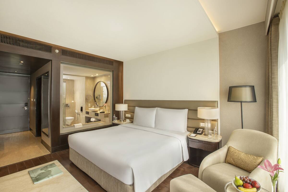 Une chambre d’hôtel avec un grand lit, un coin salon comprenant une chaise et une table, une table de chevet avec une lampe, et un miroir au-dessus du lit.