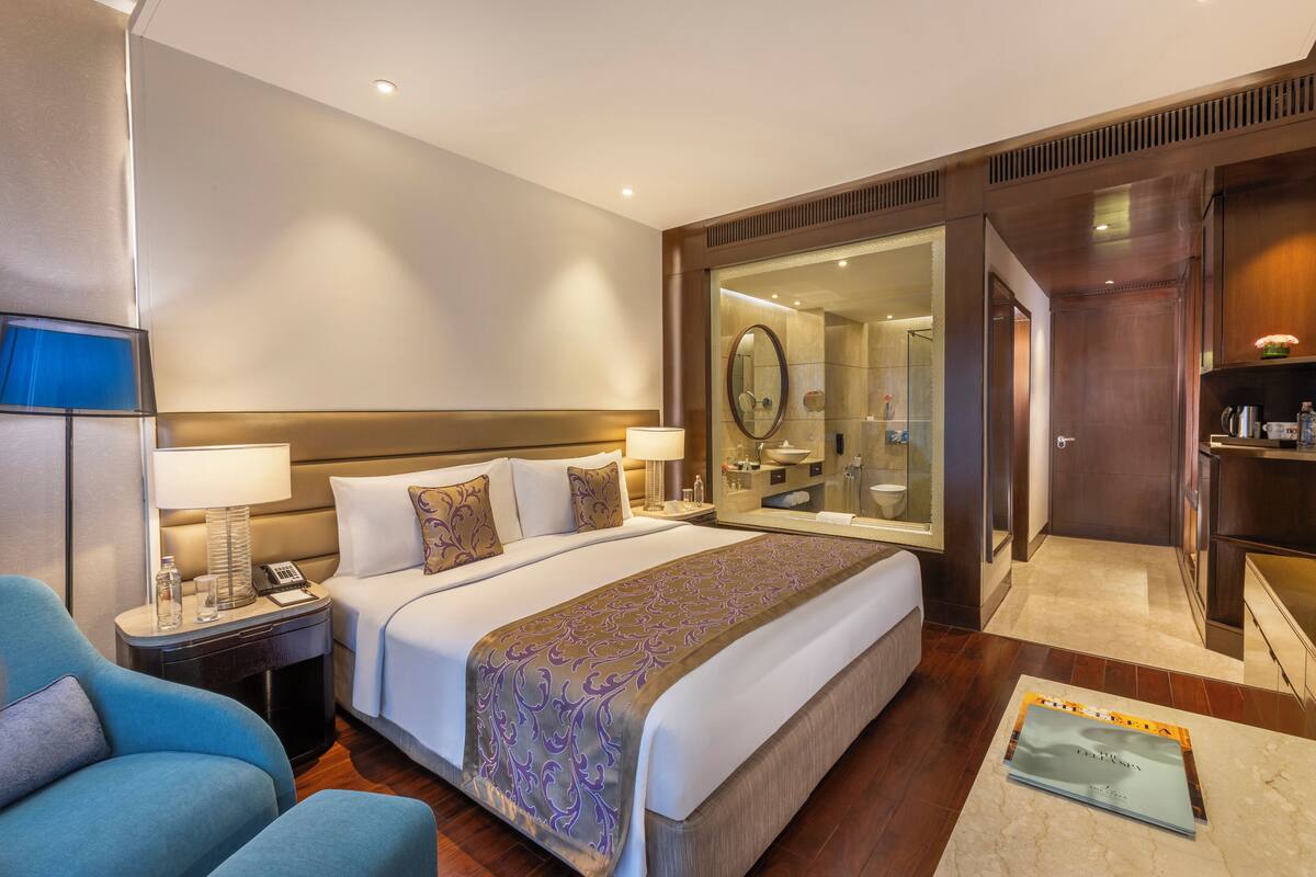 Une chambre d’hôtel avec un grand lit, une table de chevet avec une lampe, une chaise, une salle de bain avec un miroir et un petit bureau avec une lampe.