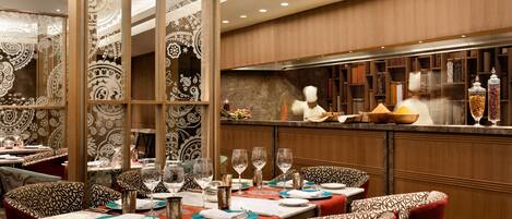 Restaurante com mesas postas para refeições, área de bar e um cozinheiro ao fundo.