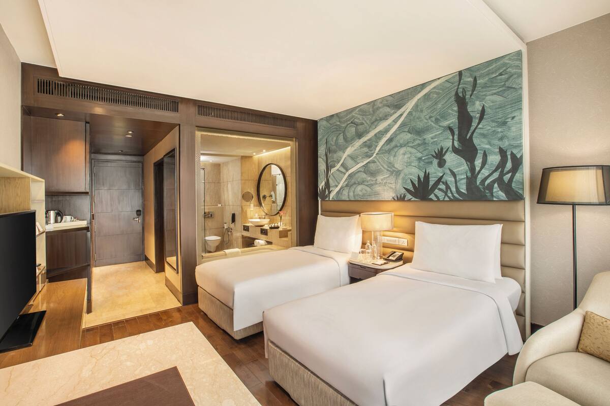 Une chambre d’hôtel avec deux lits, un bureau, une chaise, un grand tableau et une salle de bain visible à travers une porte ouverte.