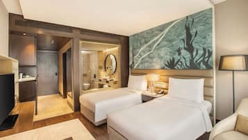 Quarto de hotel com duas camas, uma mesa, uma cadeira, um quadro grande e um banheiro visível por uma porta aberta.