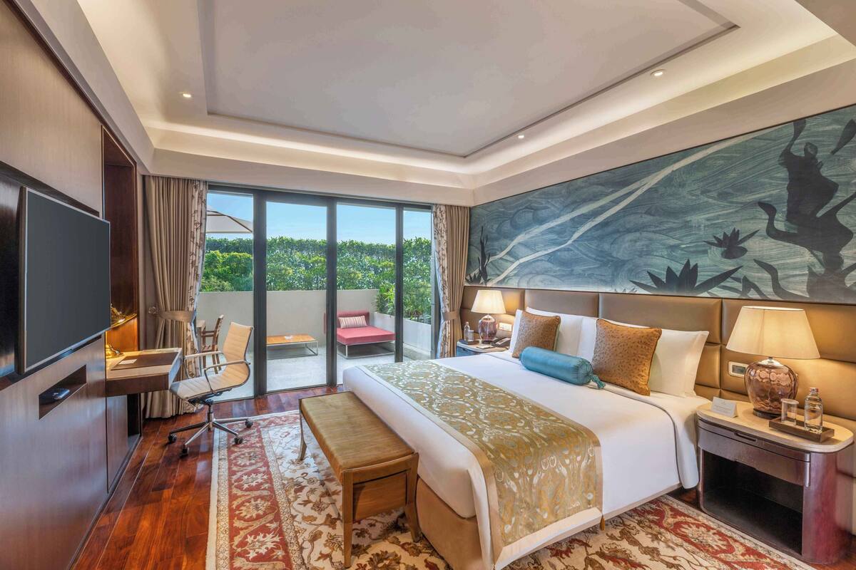 Une chambre d’hôtel spacieuse, dotée d’un grand lit, d’un bureau, d’une chaise, d’une télévision et d’un balcon offrant une vue sur la verdure.