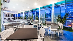 Un café moderno al aire libre con mesas y sillas, amplios ventanales con vistas a la ciudad e iluminación interior.
