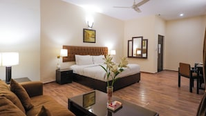 Quarto de hotel com cama, área de estar com sofá, mesa de centro, vaso com flores, ventilador de teto e espelho.