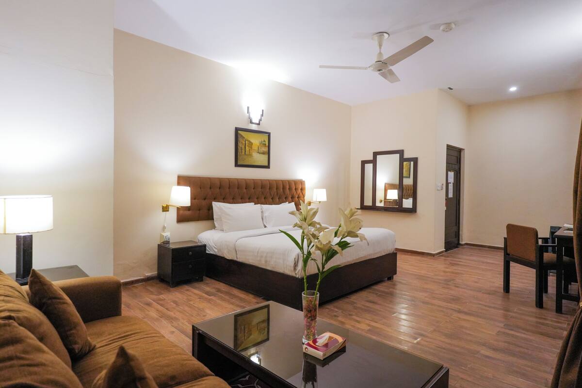 Een hotelkamer met een bed, een zithoek met een bank, een salontafel, een vaas met bloemen, een plafondventilator en een spiegel.