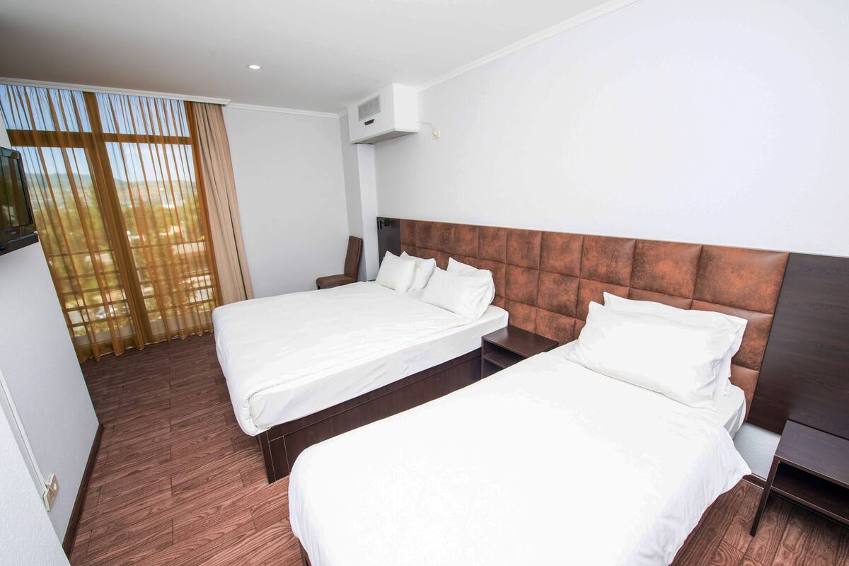 Habitación de hotel con dos camas individuales, suelo de madera, un ventanal grande con cortinas y una mesita de noche pequeña.