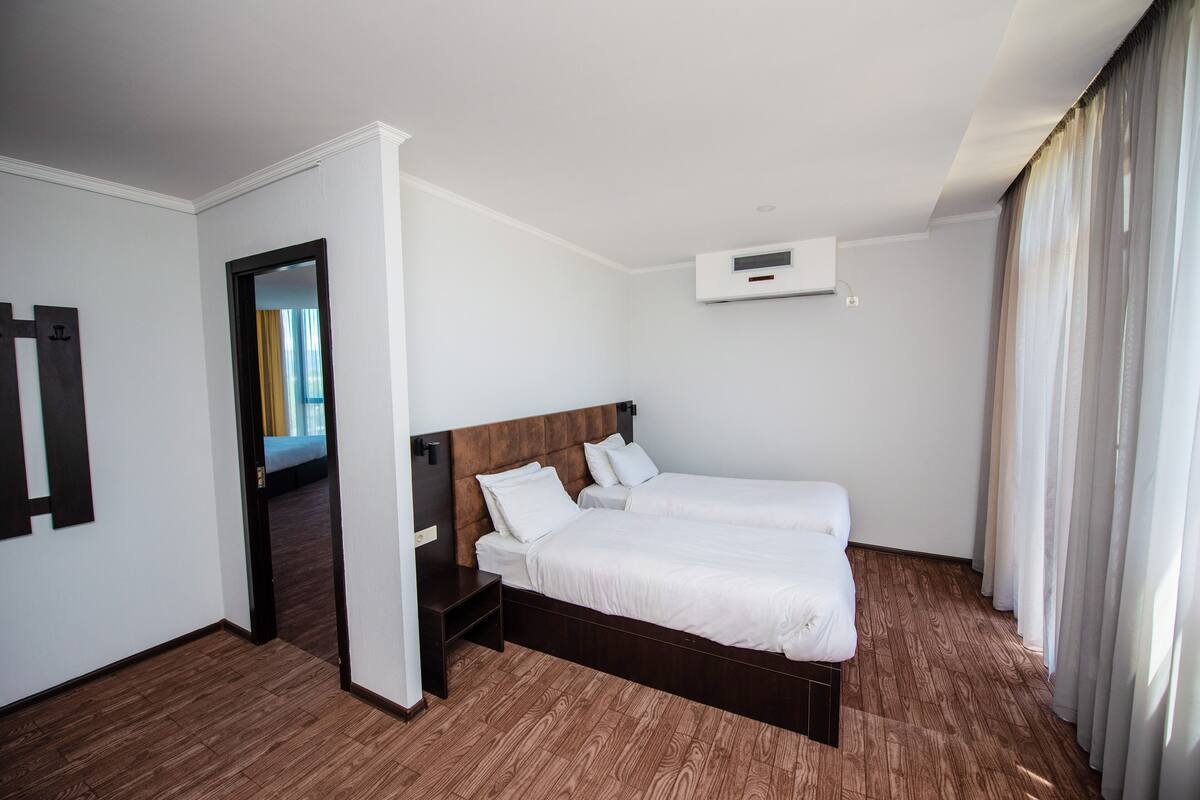 Una habitación de hotel con piso de madera, una cama con ropa blanca, una mesita de noche pequeña, un aire acondicionado instalado en la pared y una puerta que da a otra habitación.