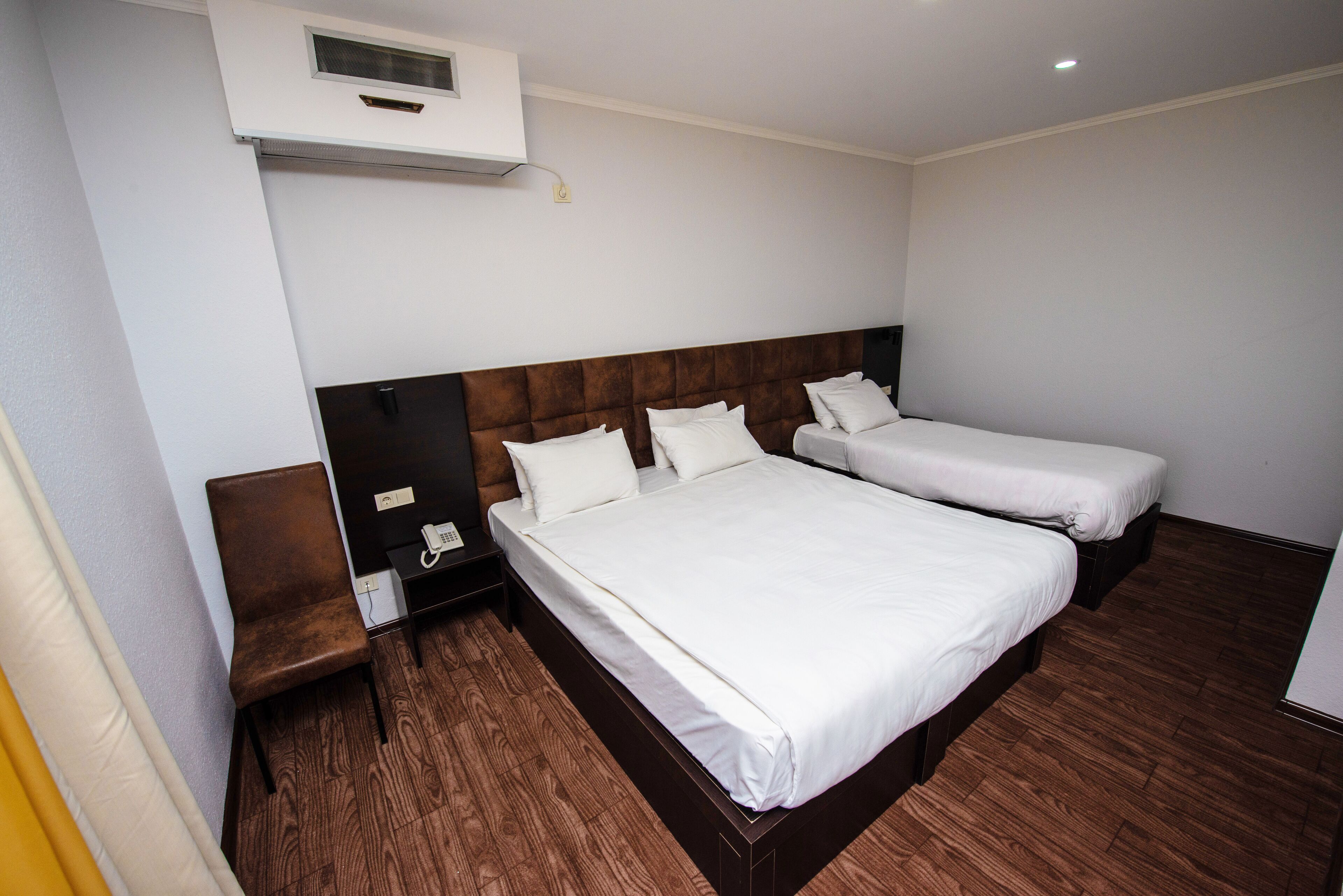 A hotel room with two beds, a wooden floor, a nightstand, and a wall-mounted air conditioner.