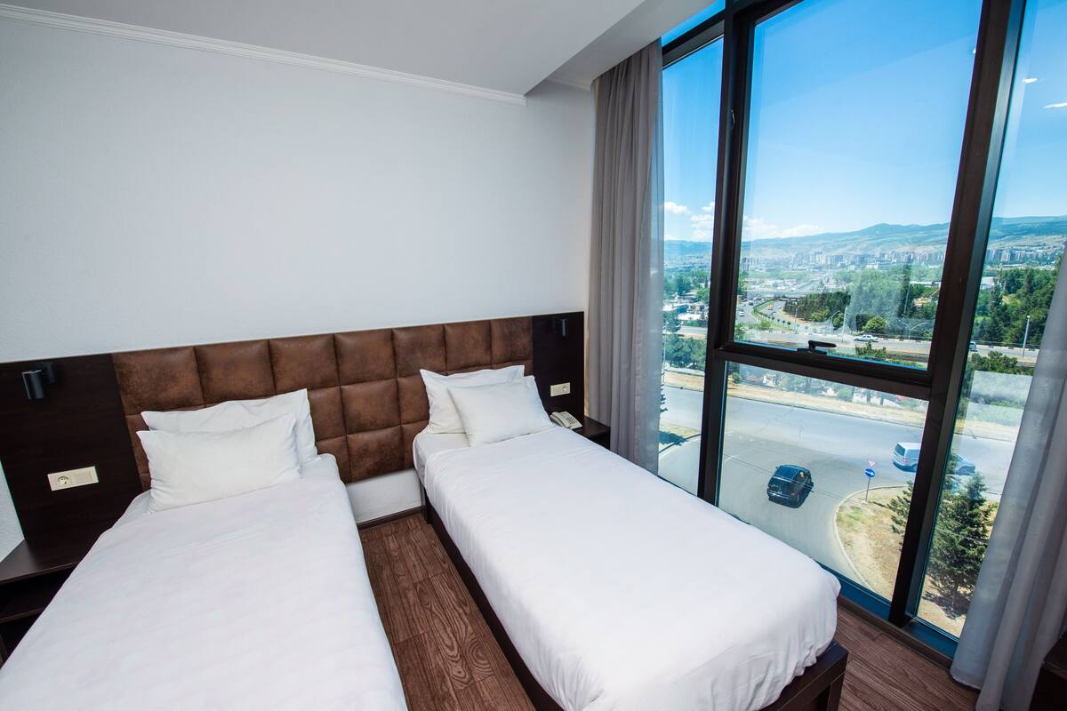 Habitación de hotel con dos camas, un ventanal grande con vistas a la ciudad y un cabecero marrón.