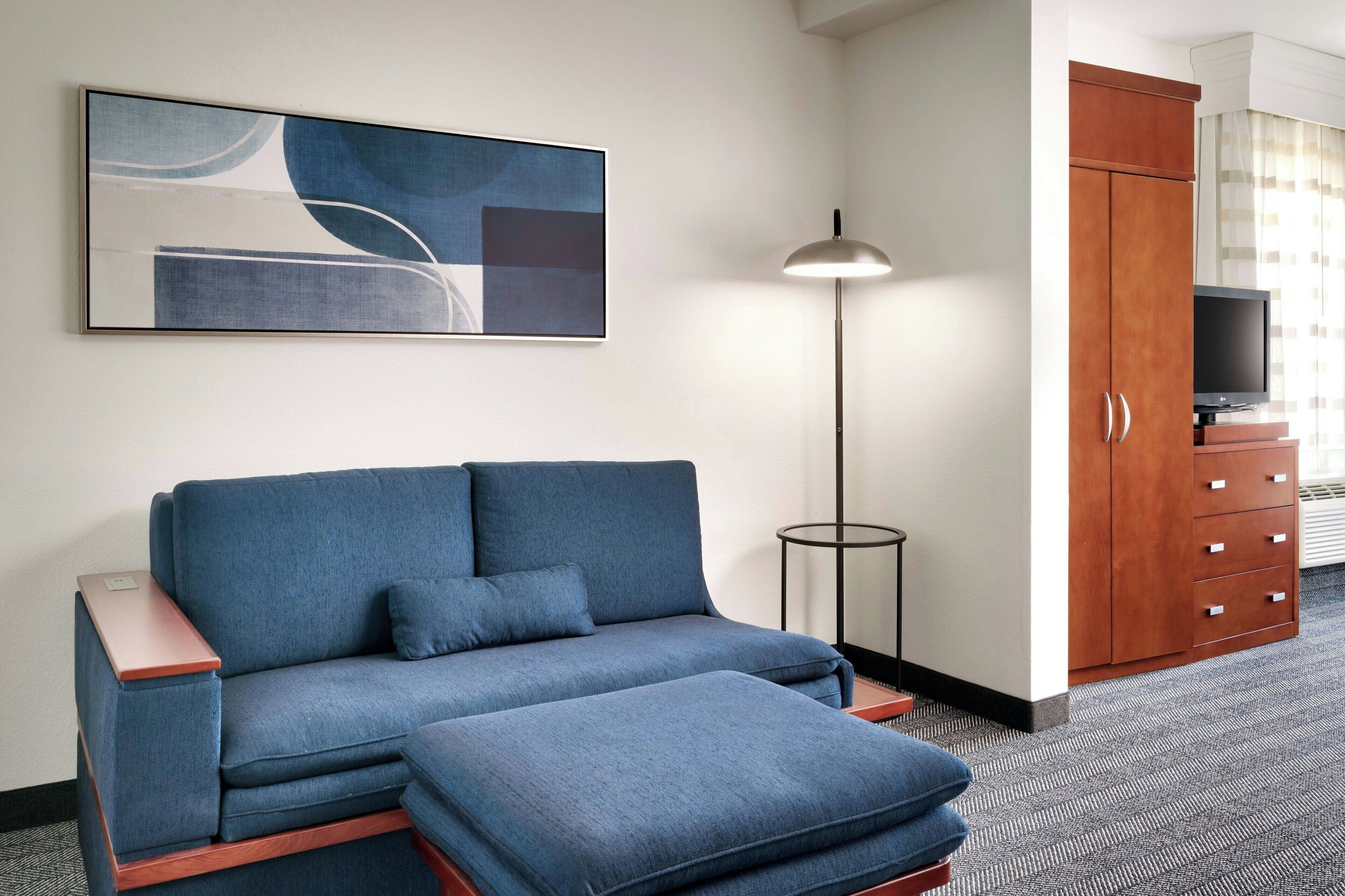 A modern hotel room with a sectional sofa, a flat-screen TV, a wooden cabinet, and a floor lamp.