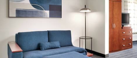 A modern hotel room with a sectional sofa, a flat-screen TV, a wooden cabinet, and a floor lamp.
