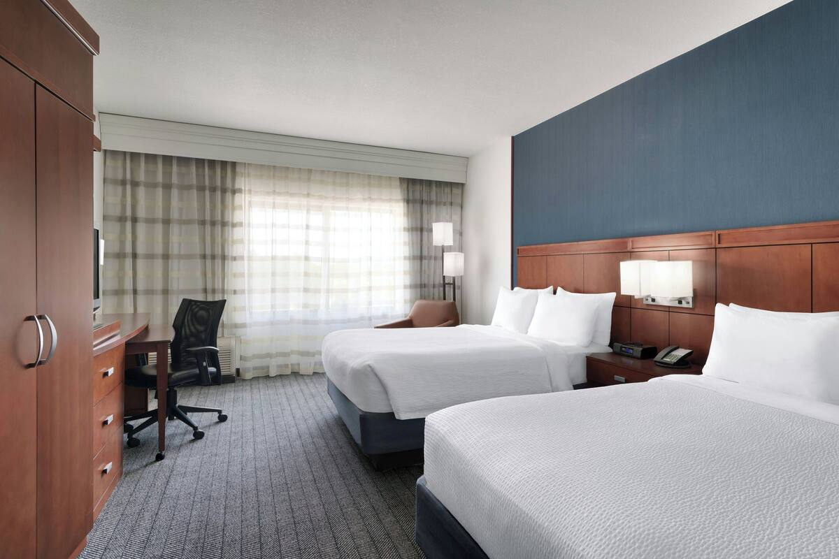 Une chambre d’hôtel avec deux lits, un bureau, une chaise, une table de chevet et une grande fenêtre avec des rideaux.