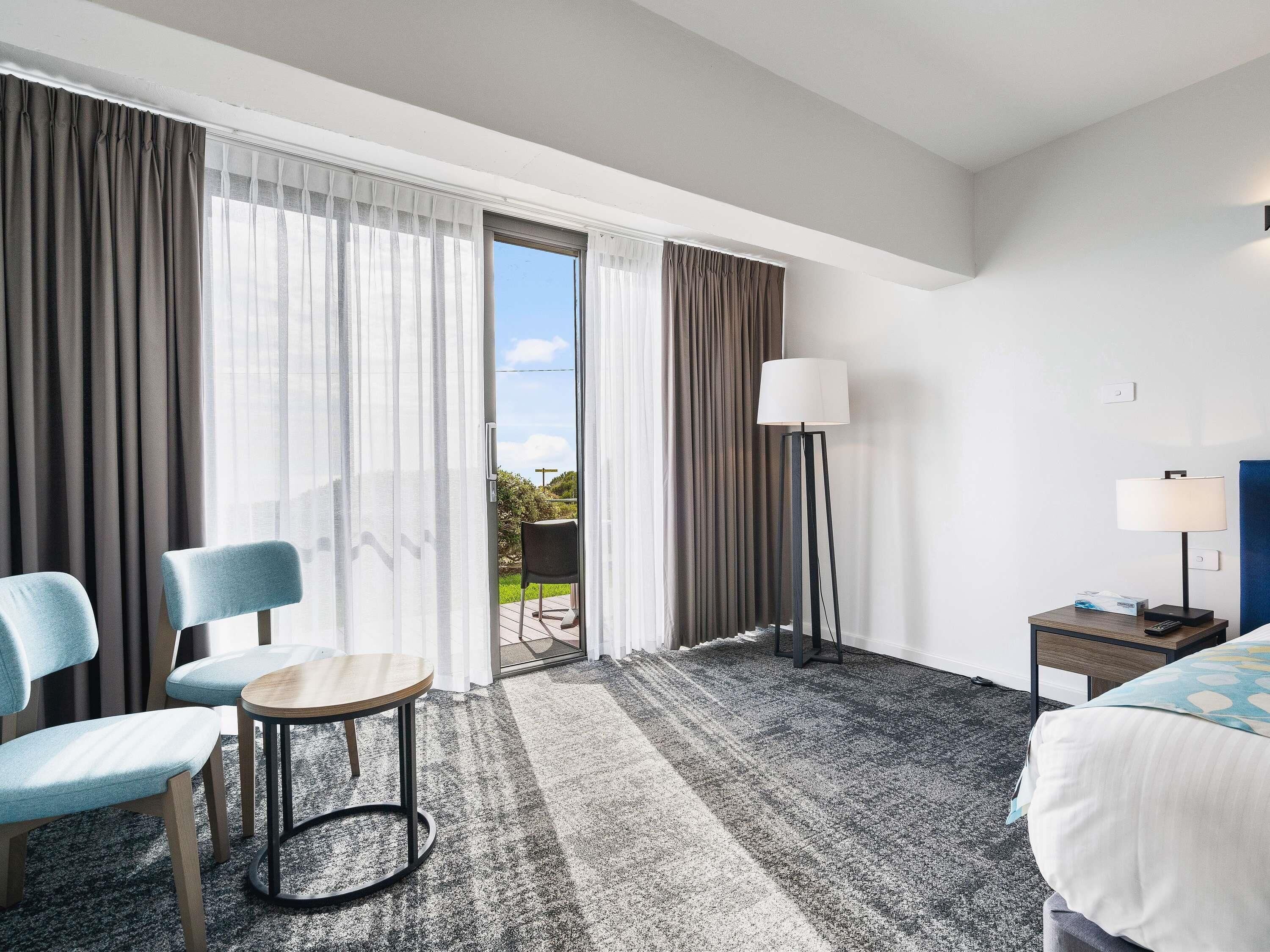 A hotel room with a bed, two chairs, a small table, a floor lamp, and a view through the door.