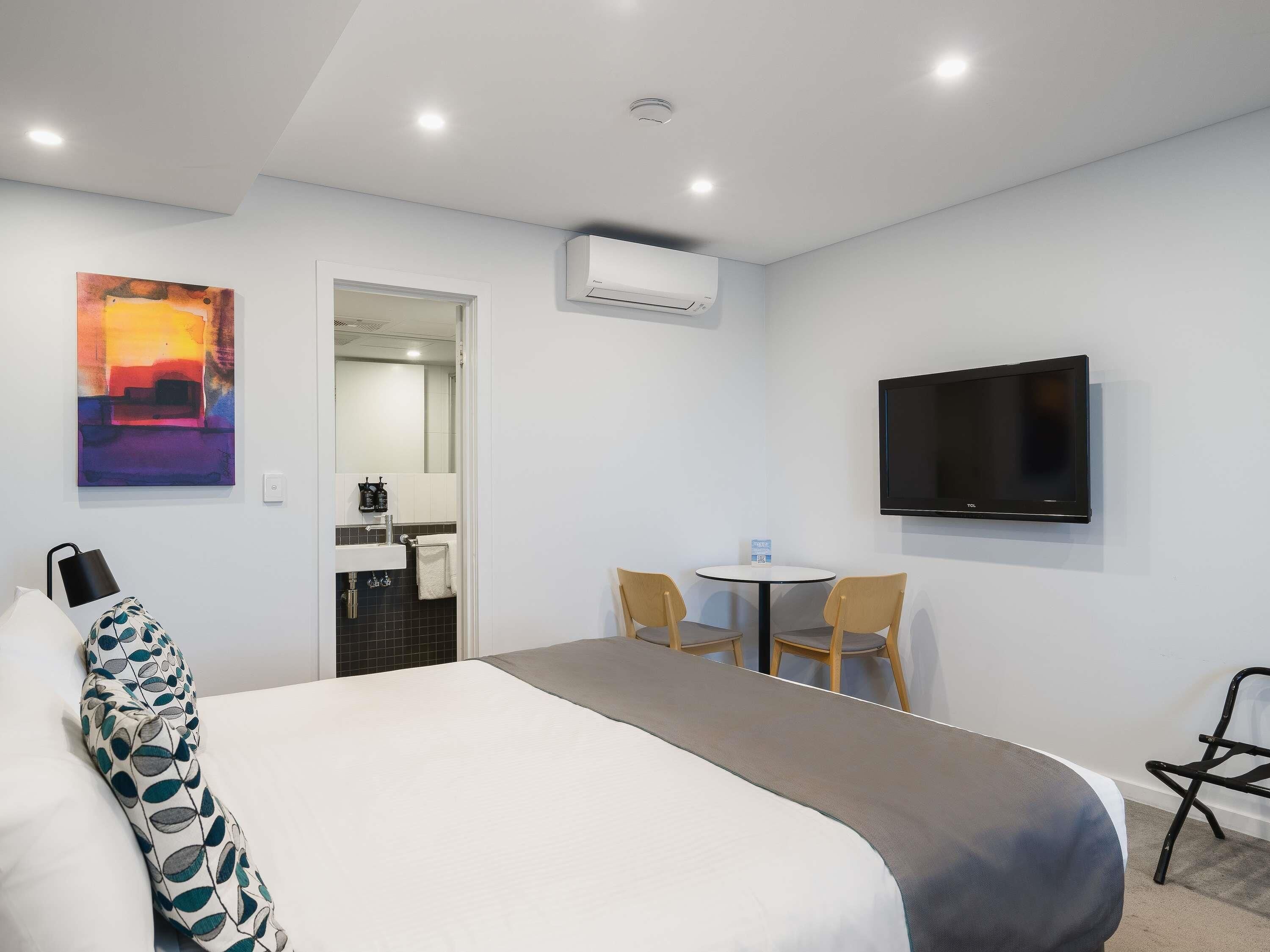 Quarto de hotel com cama, escrivaninha, cadeira, televisão, ar condicionado e um quadro na parede.