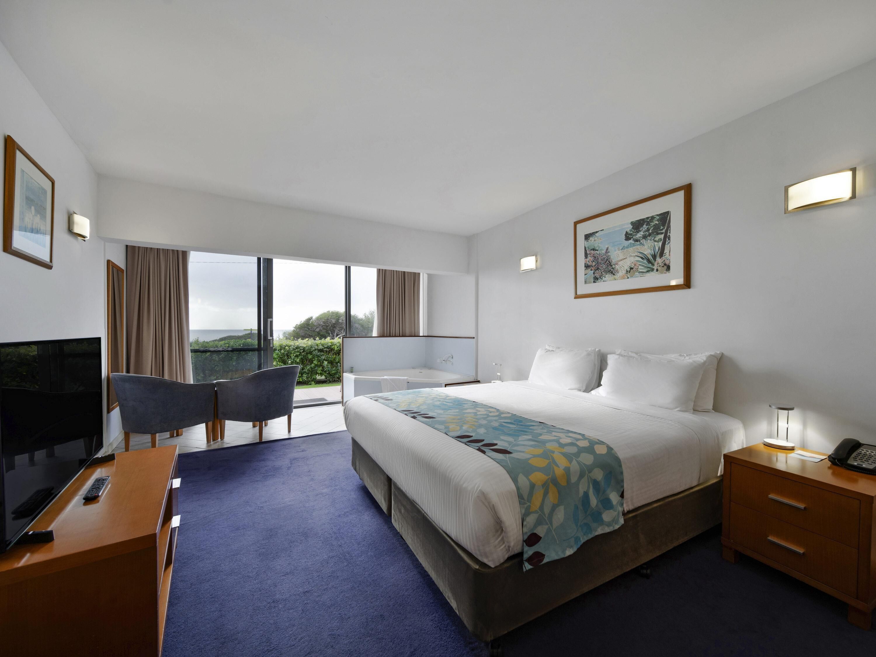 A hotel room with a bed, a desk with a TV, a chair, a bedside table with a phone, and a large window with a view.