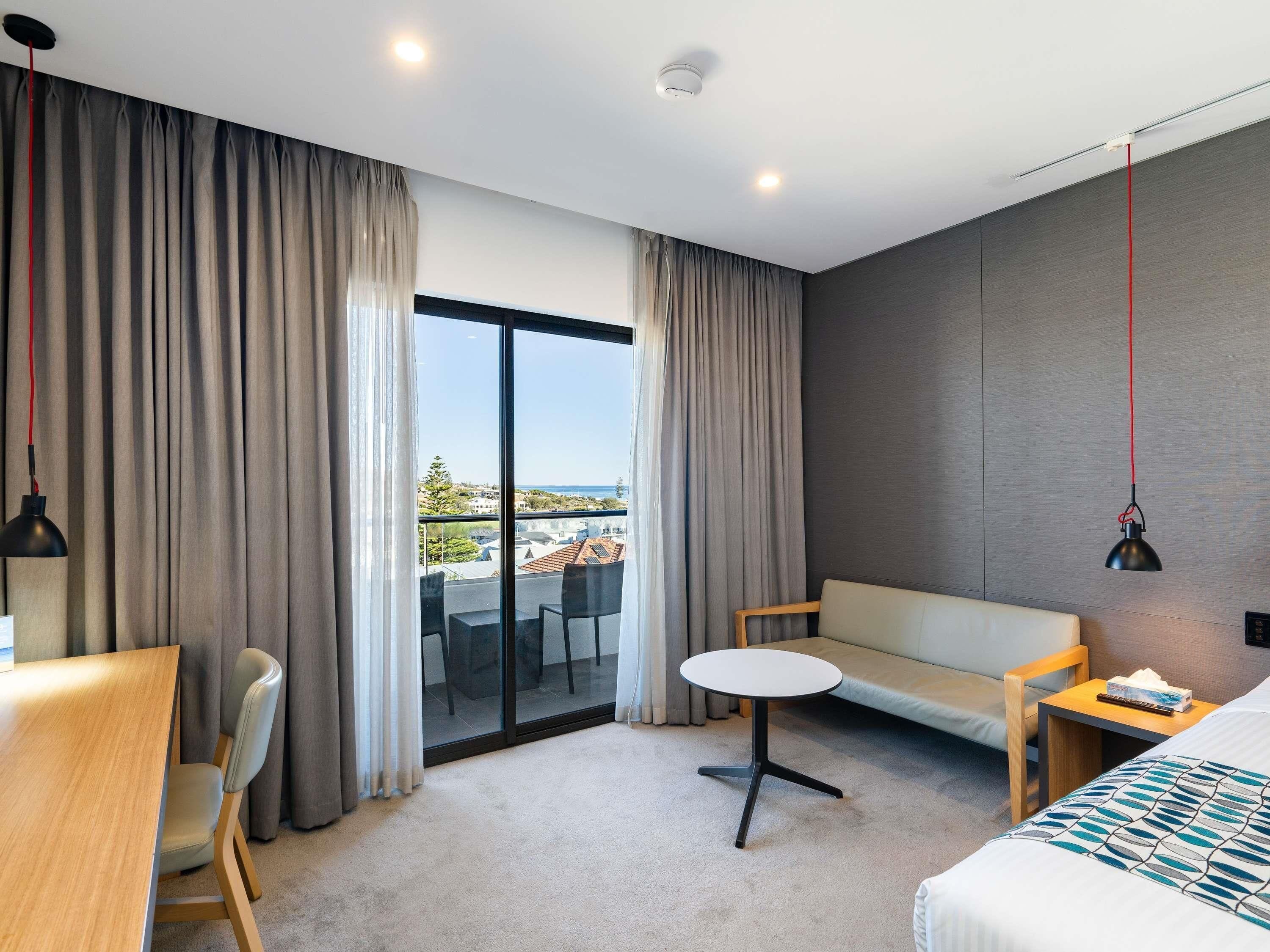A modern hotel room with a large sliding glass door, a sofa, a small round table, a desk, and a bed.