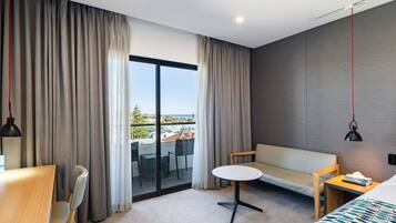 A modern hotel room with a large sliding glass door, a sofa, a small round table, a desk, and a bed.