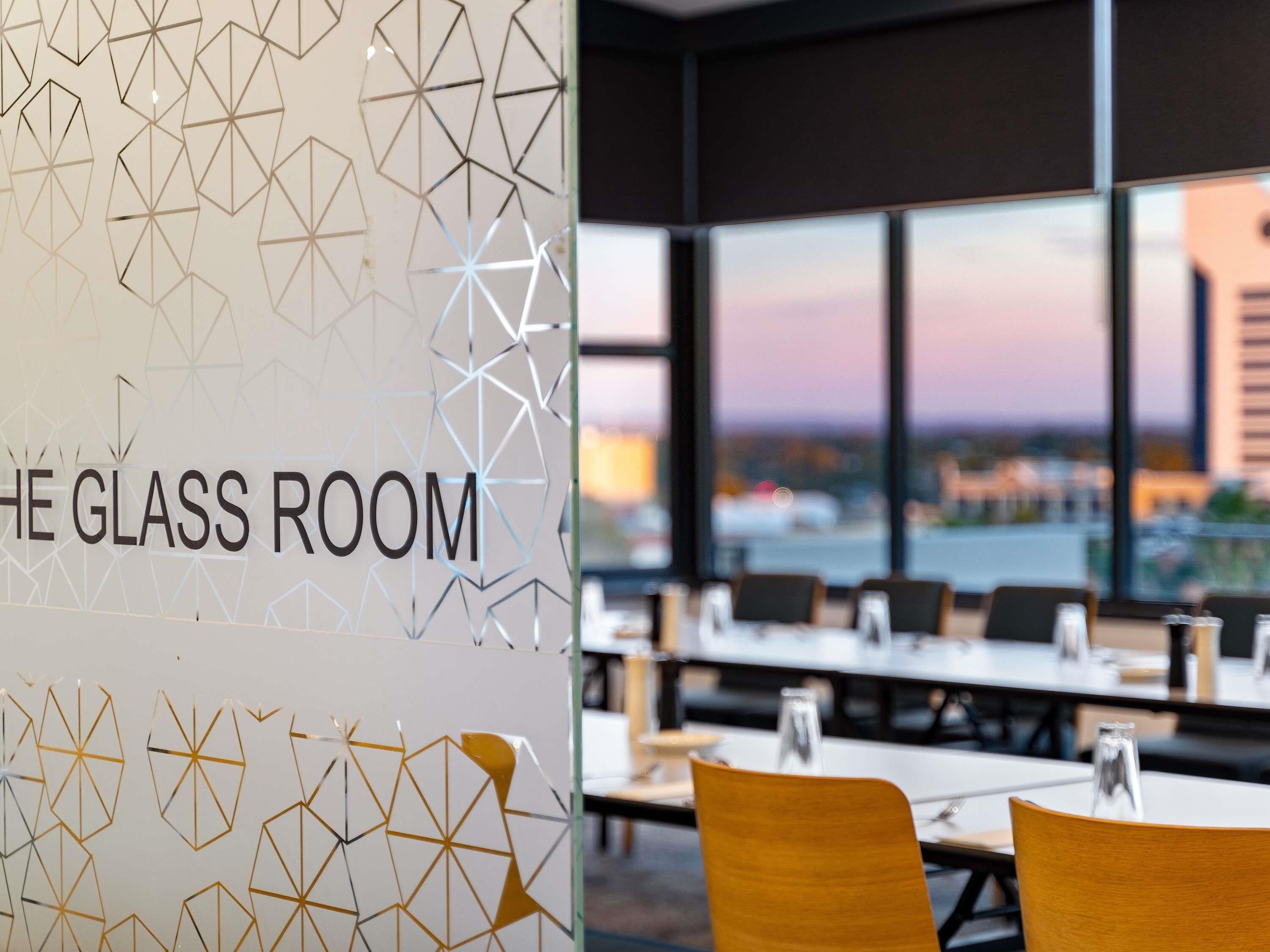 Un intérieur moderne avec un motif géométrique sur un mur, baptisé « La salle de verre », comprenant un espace repas avec des tables et des chaises, ainsi que de grandes fenêtres offrant une vue sur la ville.