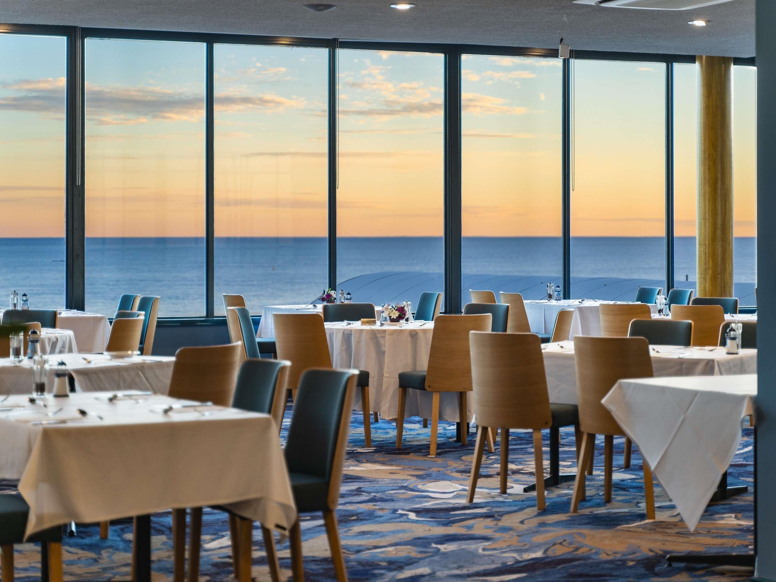 A restaurant with tables set for dining, large windows offering a view of the ocean, and a carpeted floor.