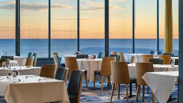 A restaurant with tables set for dining, large windows offering a view of the ocean, and a carpeted floor.