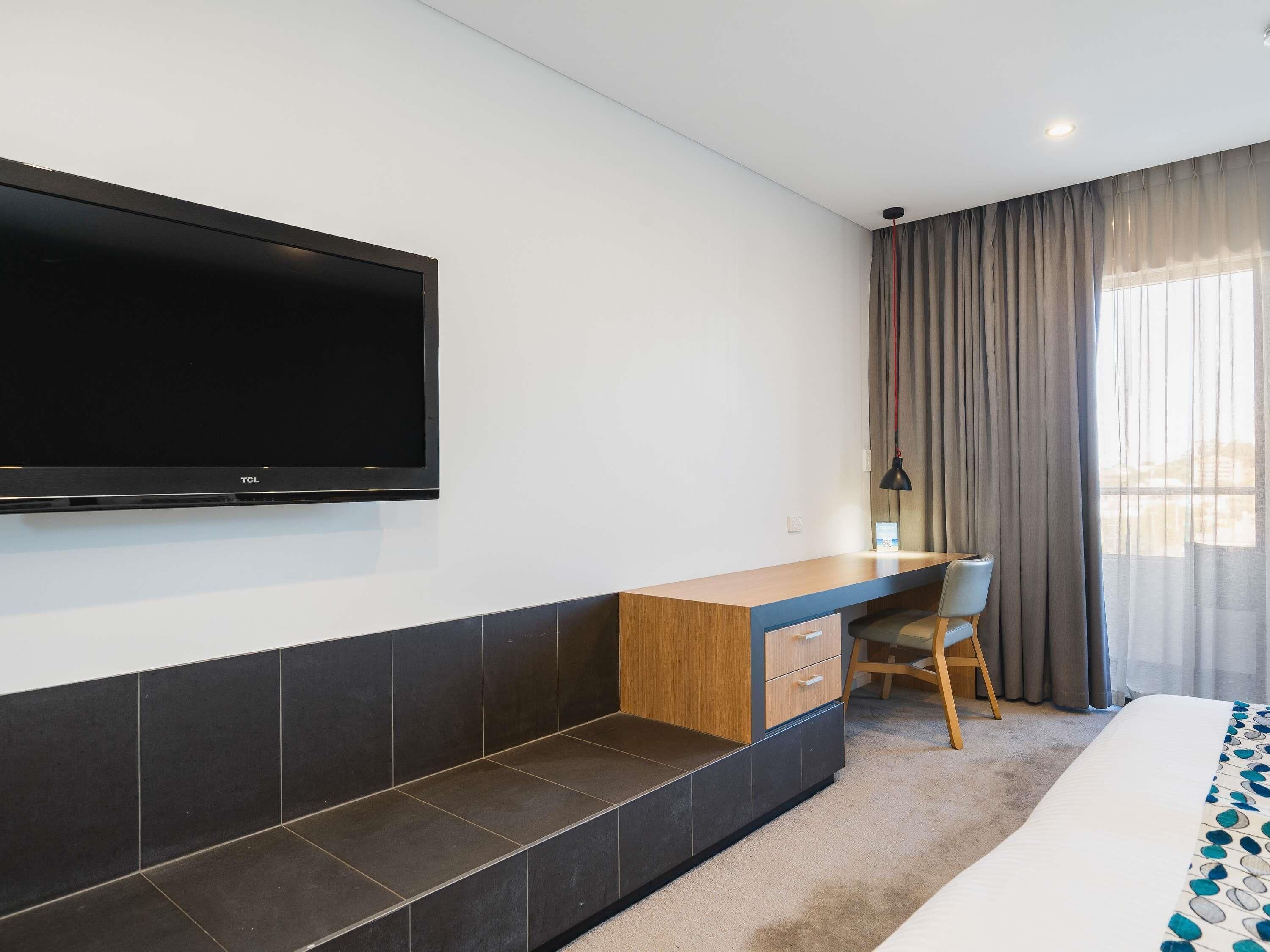 A hotel room with a flat-screen TV mounted on the wall, a wooden desk with a chair, a bed with a patterned bedspread, and a view of the city through large windows with curtains.