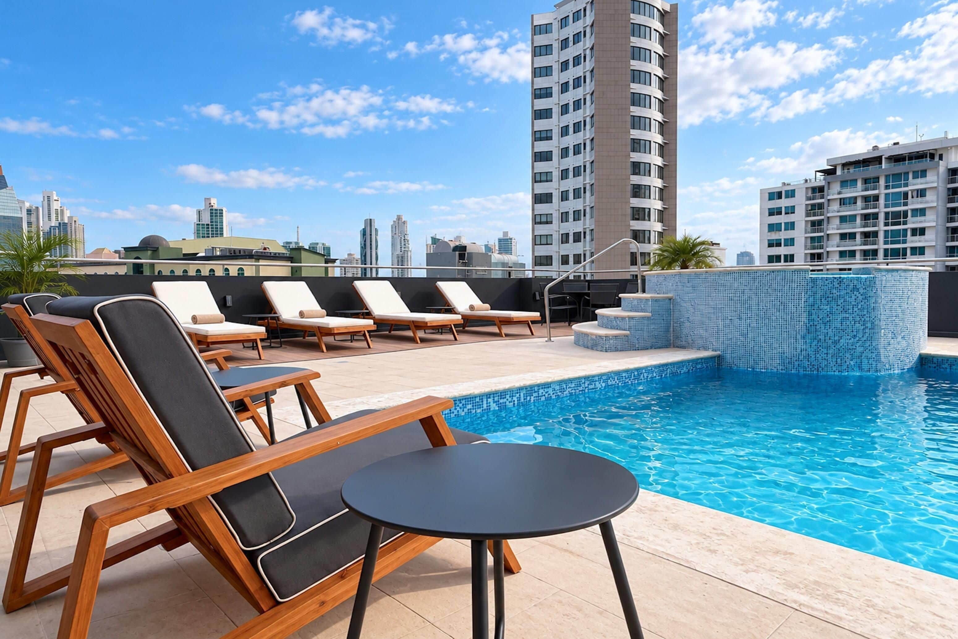 Un espace piscine aménagé sur le toit, avec des chaises longues et une petite table, entouré de grands immeubles.