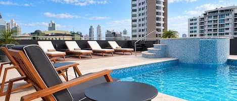 Un espace piscine aménagé sur le toit, avec des chaises longues et une petite table, entouré de grands immeubles.