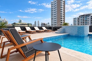 Un área de piscina en la azotea con sillones de descanso y una pequeña mesa, rodeada de edificios altos.