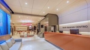 Zona de recepción de TRYP PANAMA CENTRO con diseño moderno, que incluye un mostrador de recepción, área de descanso y una pantalla grande.