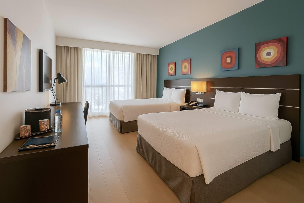 Habitación de hotel con dos camas, un escritorio con lámpara, televisión y obras de arte en las paredes.