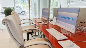 Una oficina moderna con dos escritorios, cada uno con una computadora, teclado y ratón. Hay cuatro sillas, y se ve una pequeña planta.