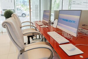 A modern office with two desks, each with a computer, keyboard, and mouse. There are four chairs, and a small plant is visible.