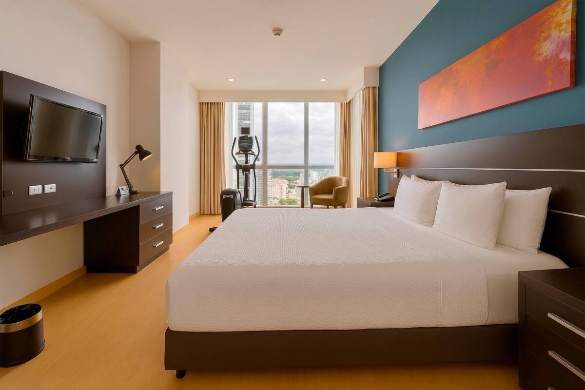 Habitación de hotel con una cama grande, televisión, escritorio con cajones, silla y ventana con vistas a edificios.
