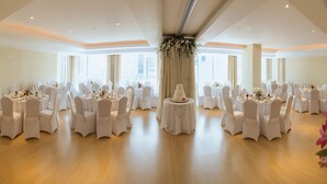 Un amplio salón de banquetes con mesas redondas y sillas blancas, ventanas grandes y una decoración floral central.