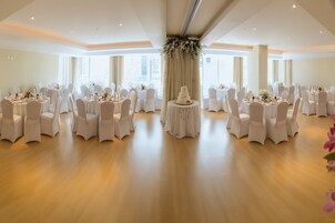 A spacious banquet hall with round tables and white chairs, large windows, and a central floral arrangement.