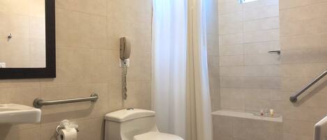 A bathroom with a toilet, a hand dryer, a trash bin, a shower with a curtain, a handrail, and a window.