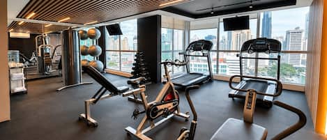 A modern gym with various exercise equipment, large windows offering city views, and a ceiling with linear lighting.