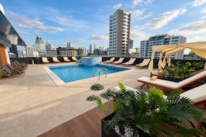 A rooftop pool with lounge chairs and potted plants, surrounded by city buildings.