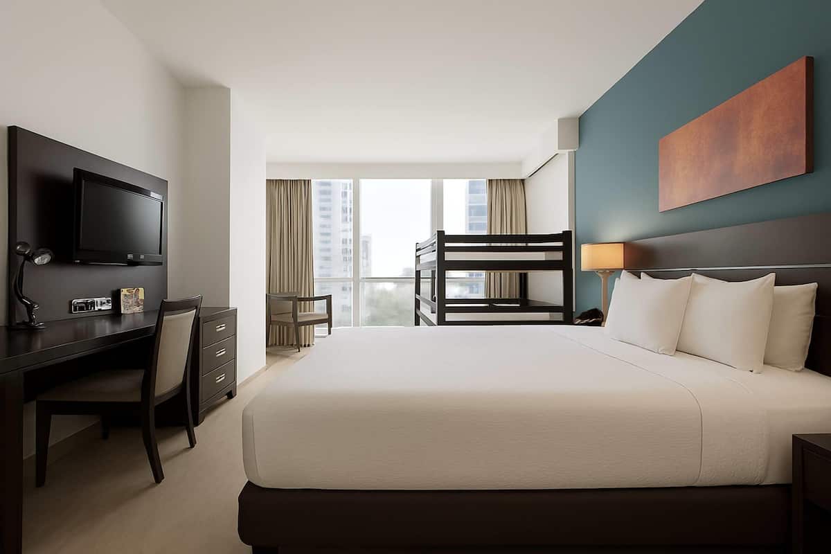 Habitación de hotel con cama, escritorio, silla, televisor, literas y vista a edificios.