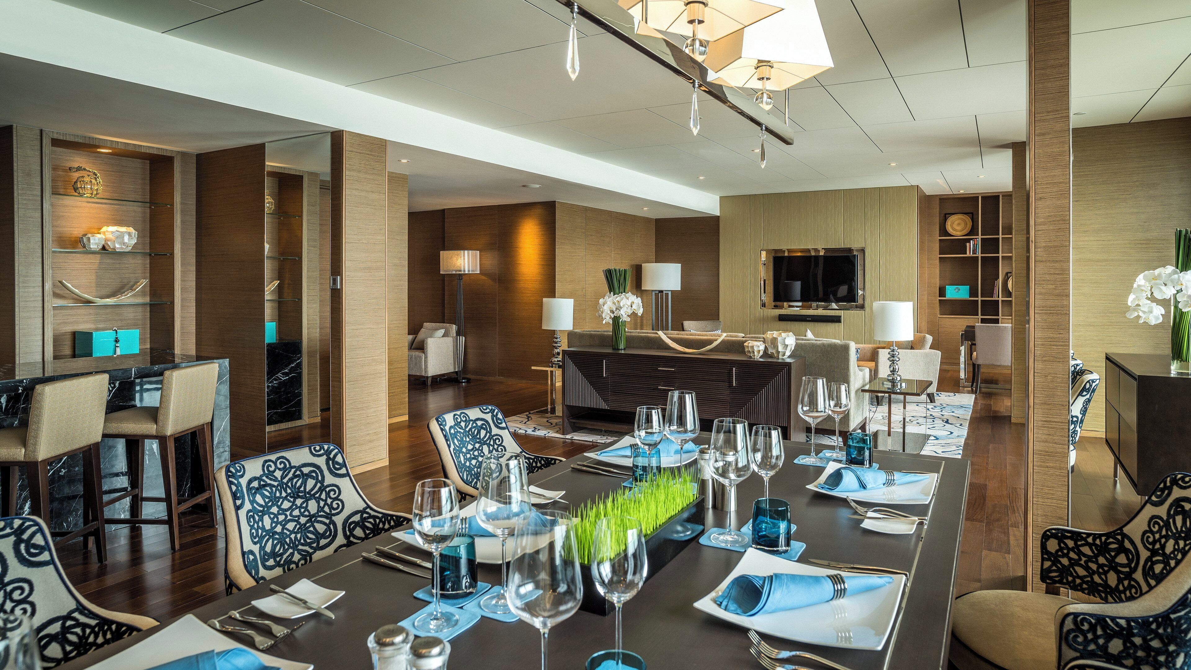 A hotel dining area with a long table set for a meal, chairs, a bar area, and a television screen.