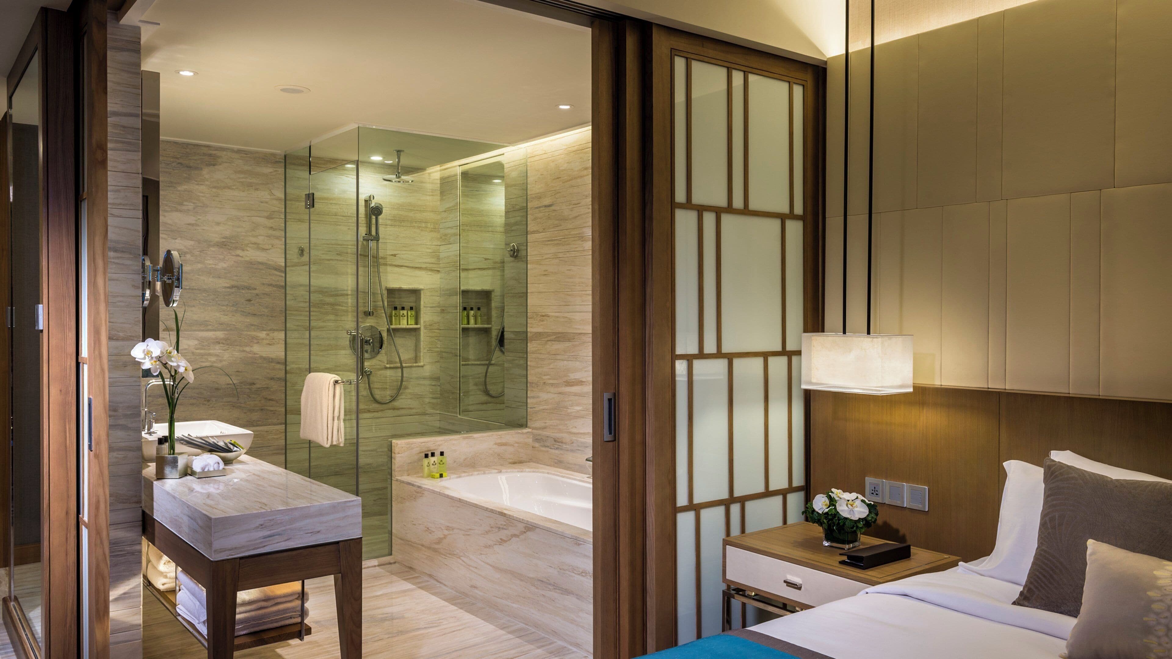 Habitación de hotel con baño que incluye ducha con mampara de vidrio, tocador de madera con espejo y cama con mesita de noche.
