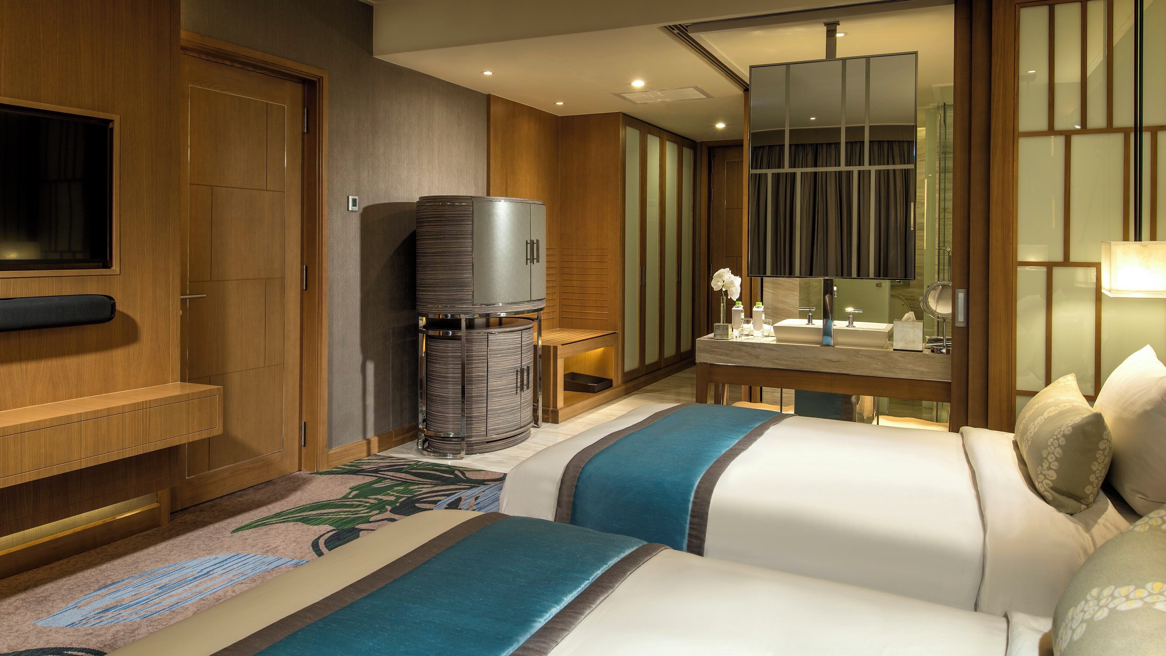 A hotel room with a bed, a TV, a minibar, a bathroom with a mirror, and a desk with a lamp.