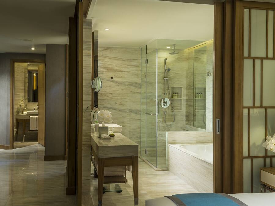 A modern hotel room with a glass-enclosed shower, a vanity with a mirror, and a bed with a blue accent pillow.