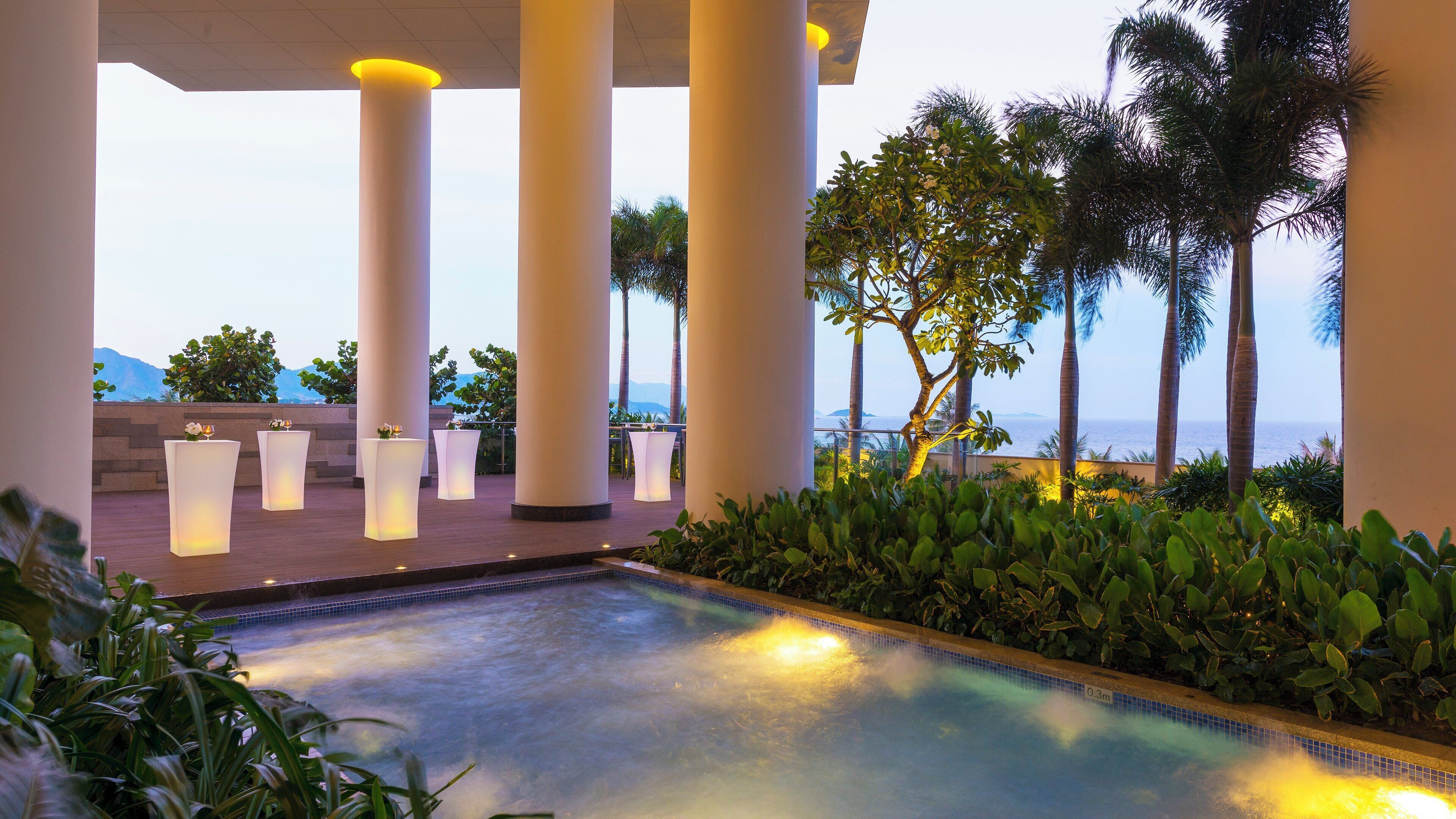 A pool area with columns, a view of the sea, and tropical plants.