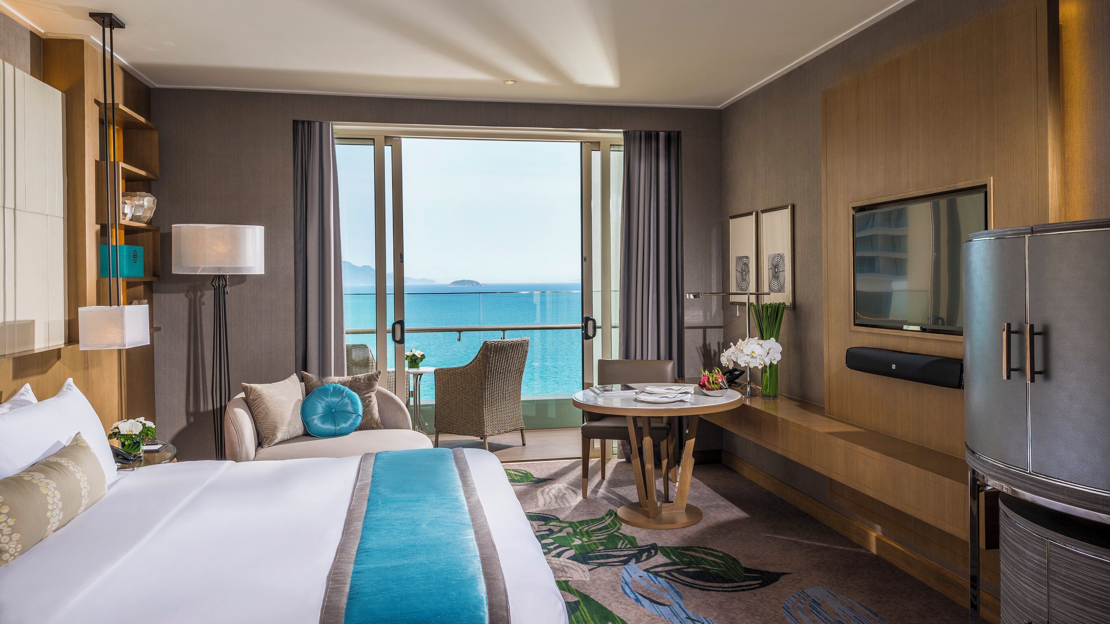 A hotel room with a bed, a dining table, a TV, and a view of the sea.