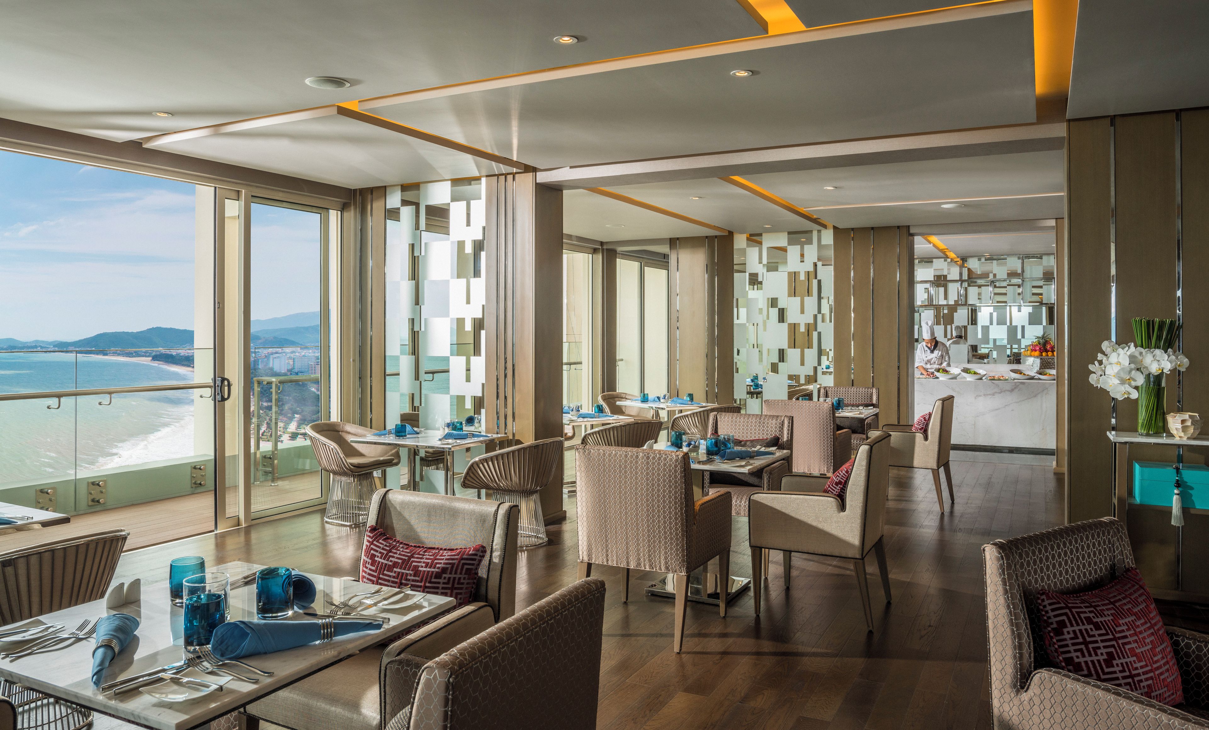 A modern dining area with tables and chairs, large windows with a view of the sea, and a kitchen visible in the background.