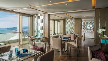A modern dining area with tables and chairs, large windows with a view of the sea, and a kitchen visible in the background.