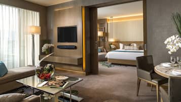 A hotel room with a large flat-screen TV, a bed with white bedding, a dining area with a table set for two, and a view of the adjacent room.