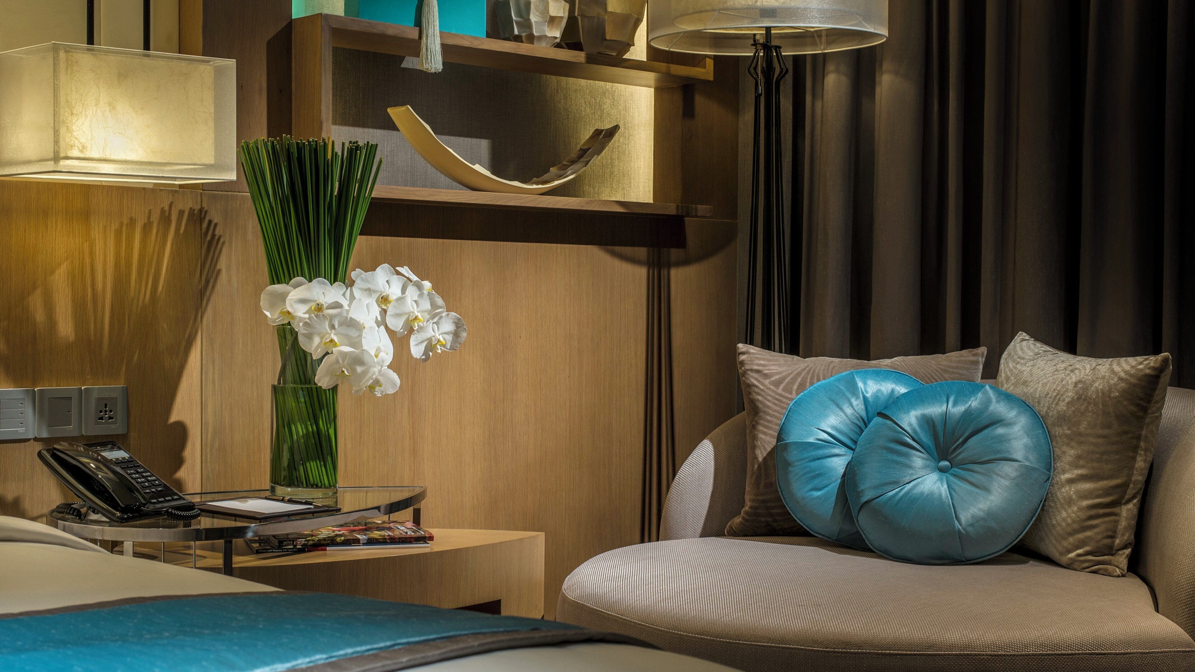 A hotel room with a bed, a bedside table with a lamp, a vase with flowers, a sofa with cushions, and a wall-mounted shelf with decorative items.