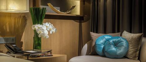 A hotel room with a bed, a bedside table with a lamp, a vase with flowers, a sofa with cushions, and a wall-mounted shelf with decorative items.